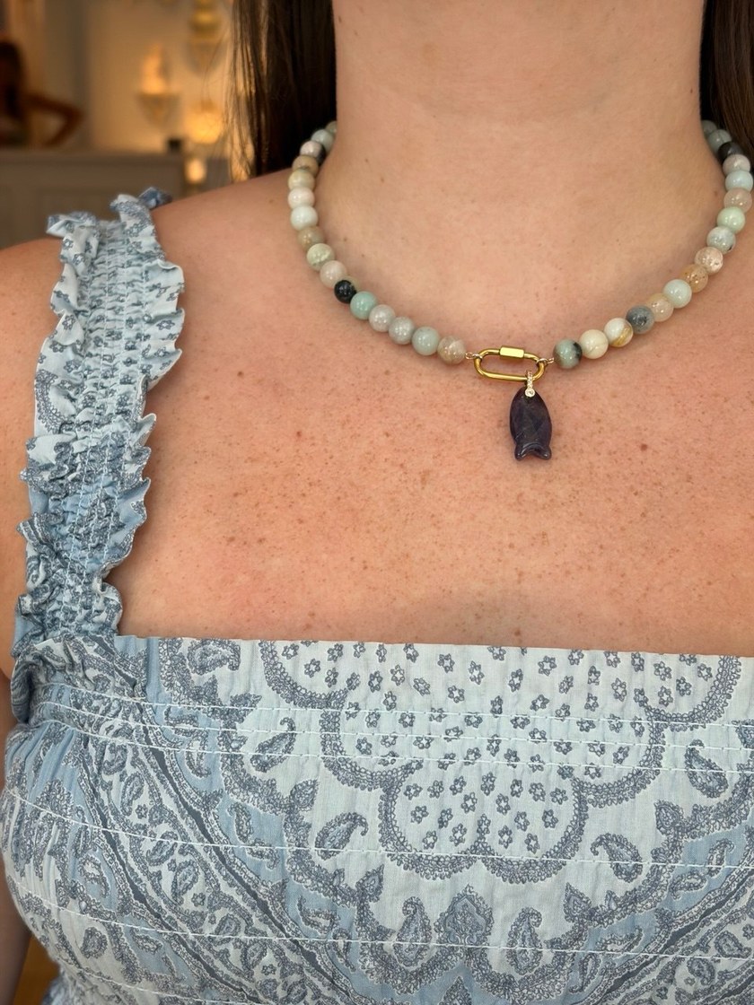 Stone strand worn at the collarbone with a gold carabiner clasp and a carved crystal fish charm hanging from it.